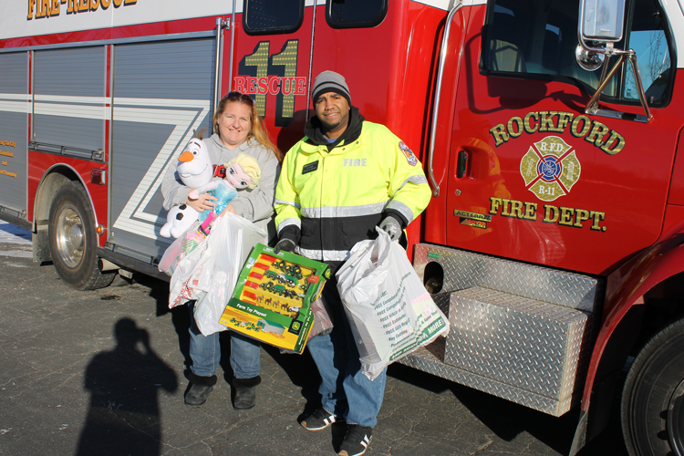 Firefighter and assistant holding donated toys with a fire truck in the background. 
